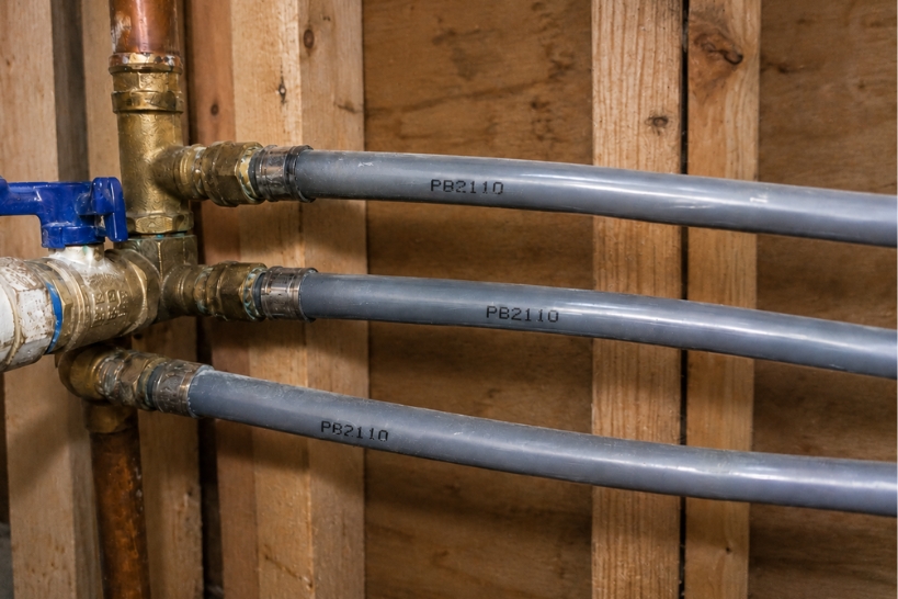 Gray polybutylene plumbing pipes connected to copper piping inside a wood-framed crawl space.